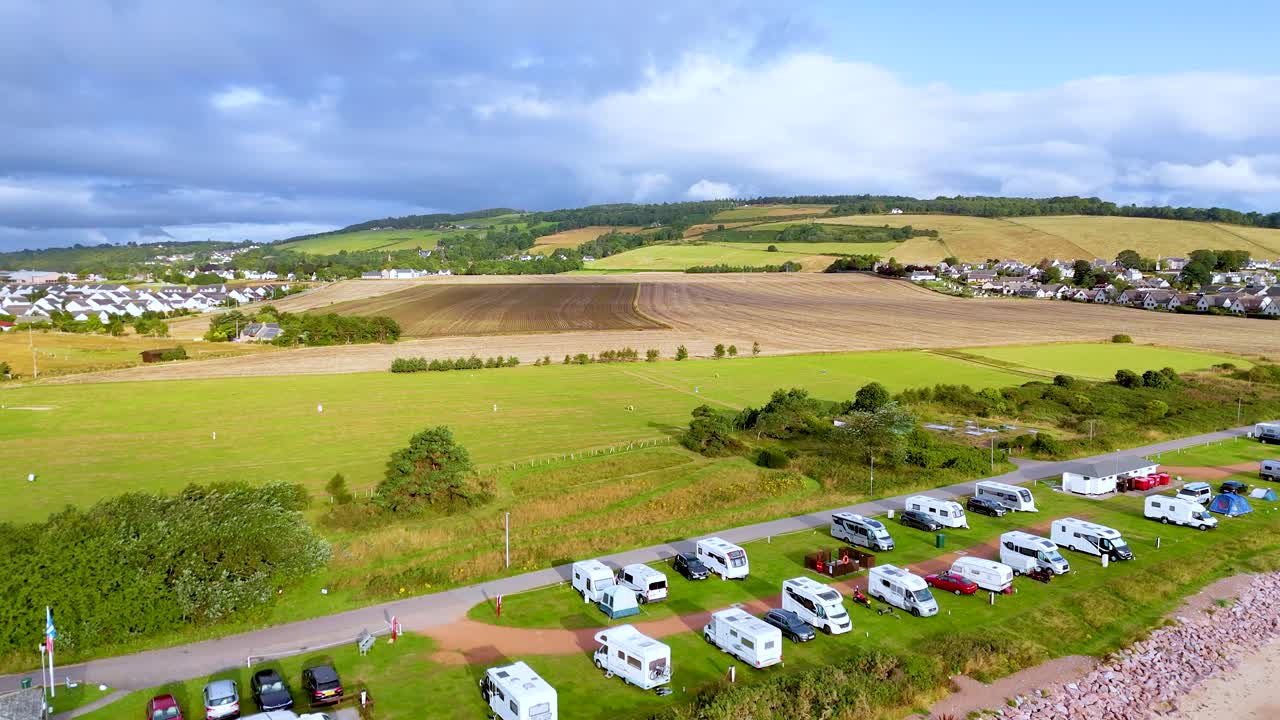 Aerial drone footage glides over rural caravan campsite, grassy fields, rolling hills, and parked vehicles