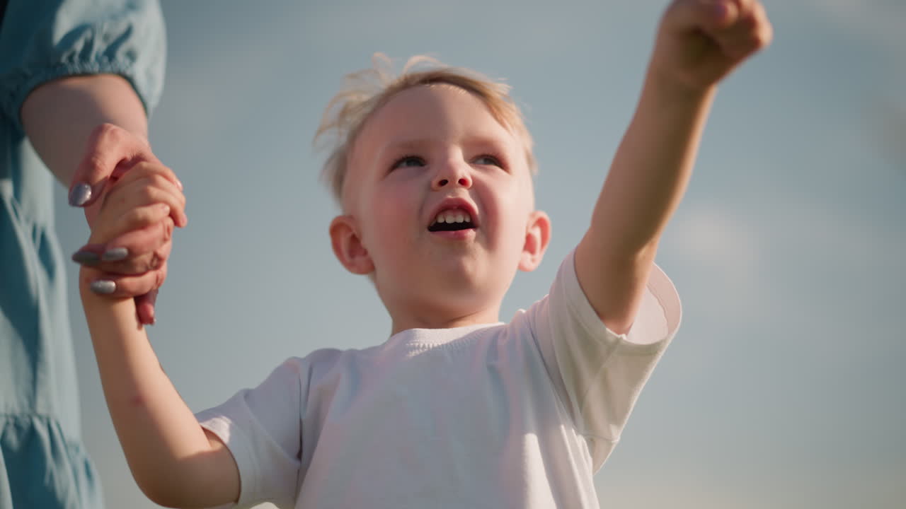 A joyful little boy in a white shirt smiles as he holds his mother s hand and points enthusiastically towards the sky. The mother is partially visible