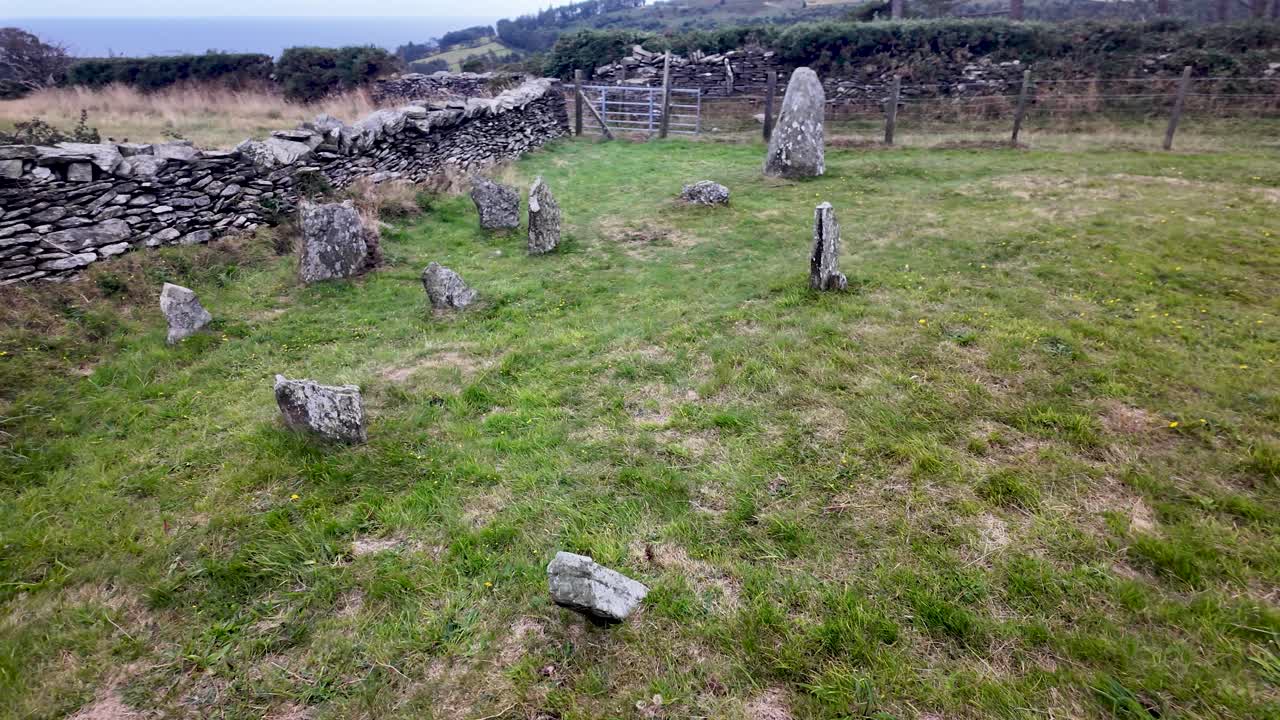 Ancient stone circle standing in a grassy field with a stone wall and distant sea