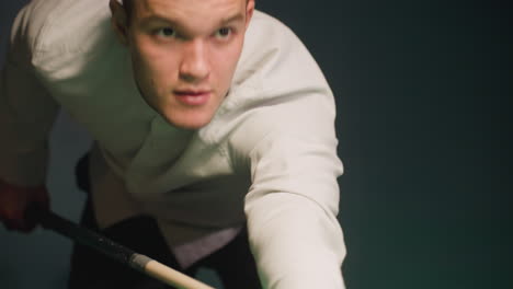 player in white shirt leans over green pool table, gripping cue stick with intense focus. eyes locked on cue ball as he takes shot, demonstrating precision, concentration