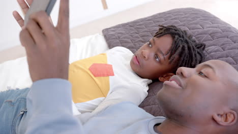 Happy-african-american-father-and-son-lying-on-bed-using-tablet-at-home,-slow-motion