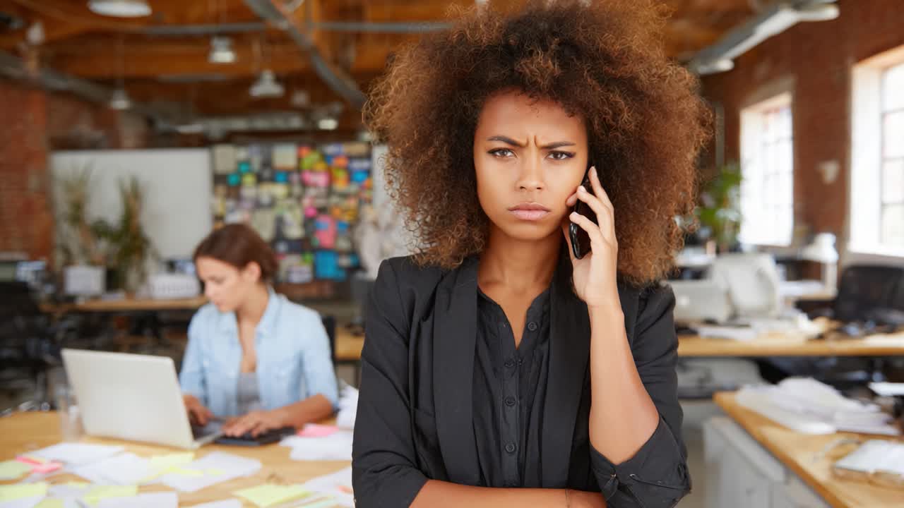 A Woman in a Modern Workspace Faces a Challenge While on the Phone, Exhibiting a Serious Expression Amidst a Busy Office Environment with Colleagues in the Background