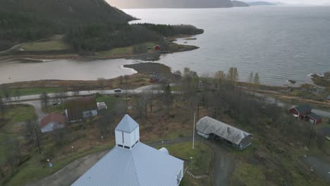 A-white-church-in-sorreisa,-norway,-near-a-fjord-with-a-cloudy-sky,-aerial-view