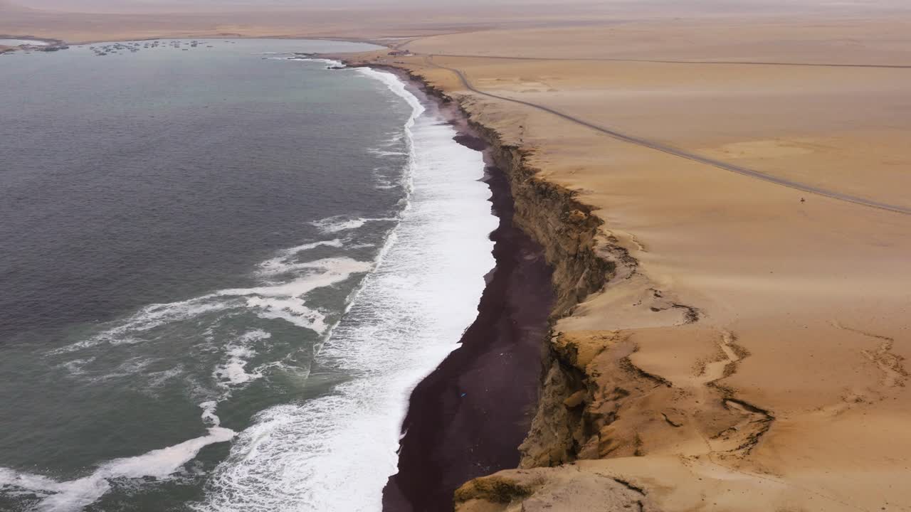 Aerial establishing of steady surf rolling against pale sand as cliffs extend into the Paracas bay
