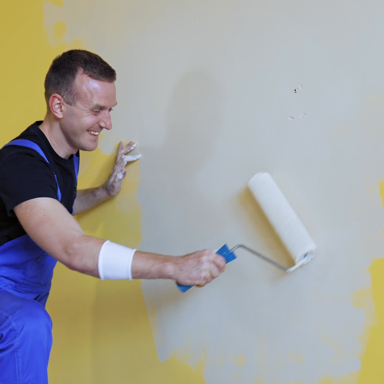 Positive worker doing renovation in the room. Smiling builder painting wall in the light color with a paint roller. Room makeover