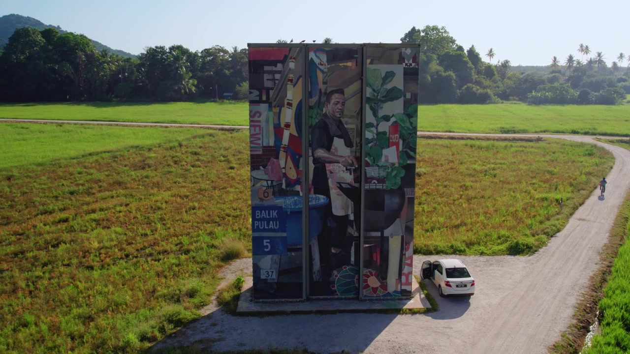 L'orbita del drone rivela l'arte dei container di Balik Pulau che si erge in modo prominente tra le verdi risaie, mentre i veicoli percorrono una stretta strada di campagna nella Penang rurale.