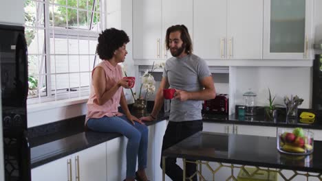 Mixed-race-couple-holding-coffee-cup-talking-to-each-other-in-the-kitchen-at-home