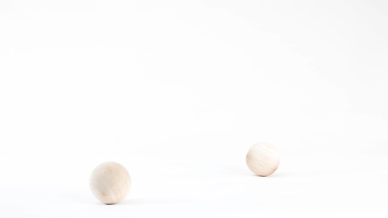 Two unpainted wooden balls fall, bounce, and roll across a bright white studio surface