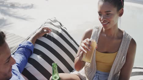 happy diverse couple talking and drinking beers sitting on beach sun deck, in slow motion