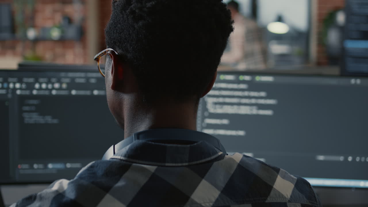 Portrait of african american programer typing on laptop sitting at desk with multiple screens parsing code