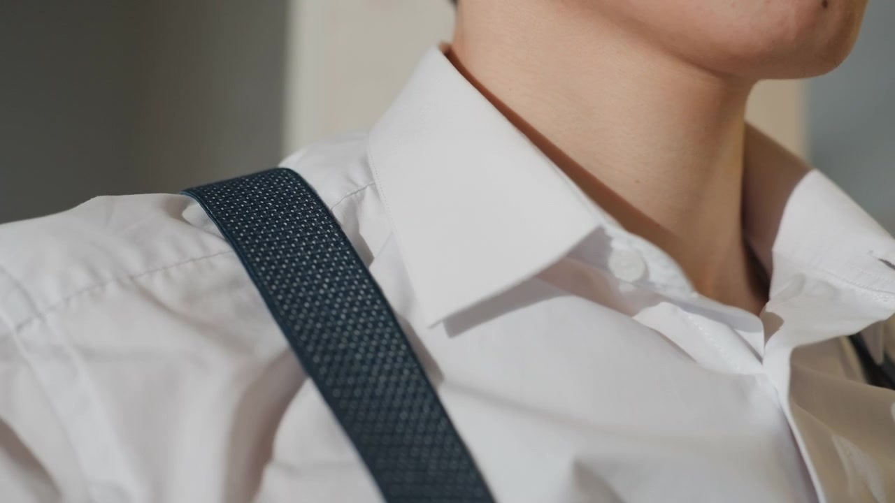 Uomo asiatico che si sistema il colletto della camicia bianca, primo piano del busto e delle mani che abbottonano i polsini mentre allaccia le bretelle blu navy, luce calda interna che proietta un'ombra morbida sulla mascella, atmosfera calma e concentrata, uomo d'affari.