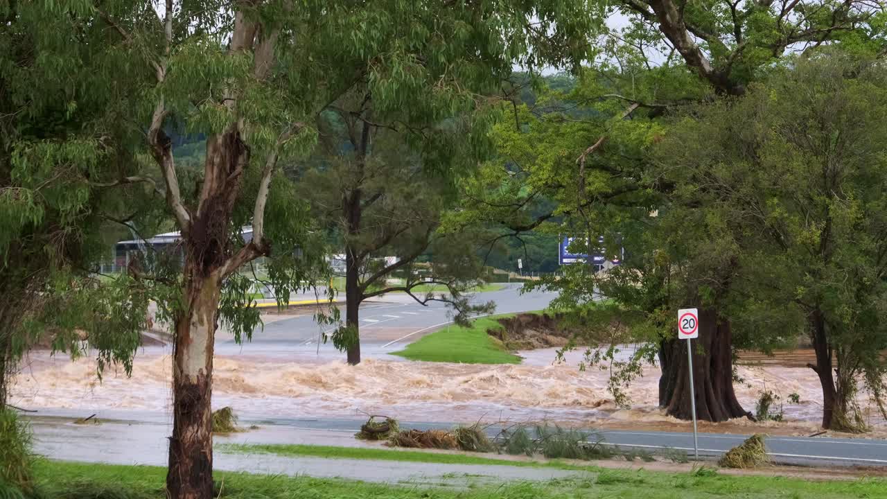 Expansive view of a flooded river bursting its banks on the Gold Coast in Queensland, Australia. Brown, churning water flows forcefully through the green riverbanks.