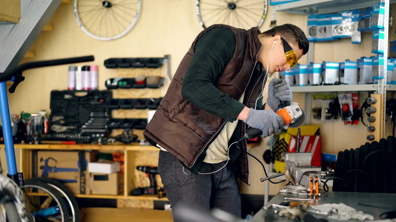 mecánico concentrado está aserrando piezas metálicas con sierra circular eléctrica de pie en la mesa de trabajo en un taller acogedor. bicicletas, piezas de repuesto y equipos son visibles.