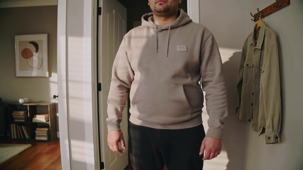 Uomo adulto in piedi che indossa una felpa con cappuccio beige mentre la telecamera si muove verso l'interno all'ingresso, mostrando l'appendiabiti.