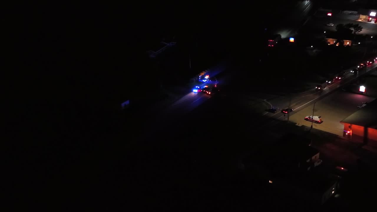 Aerial approaching shot of night scene at car accident in american town. Lighting shops and stores in suburbia. Zoom shot. Flashing lights of emergency vehicles