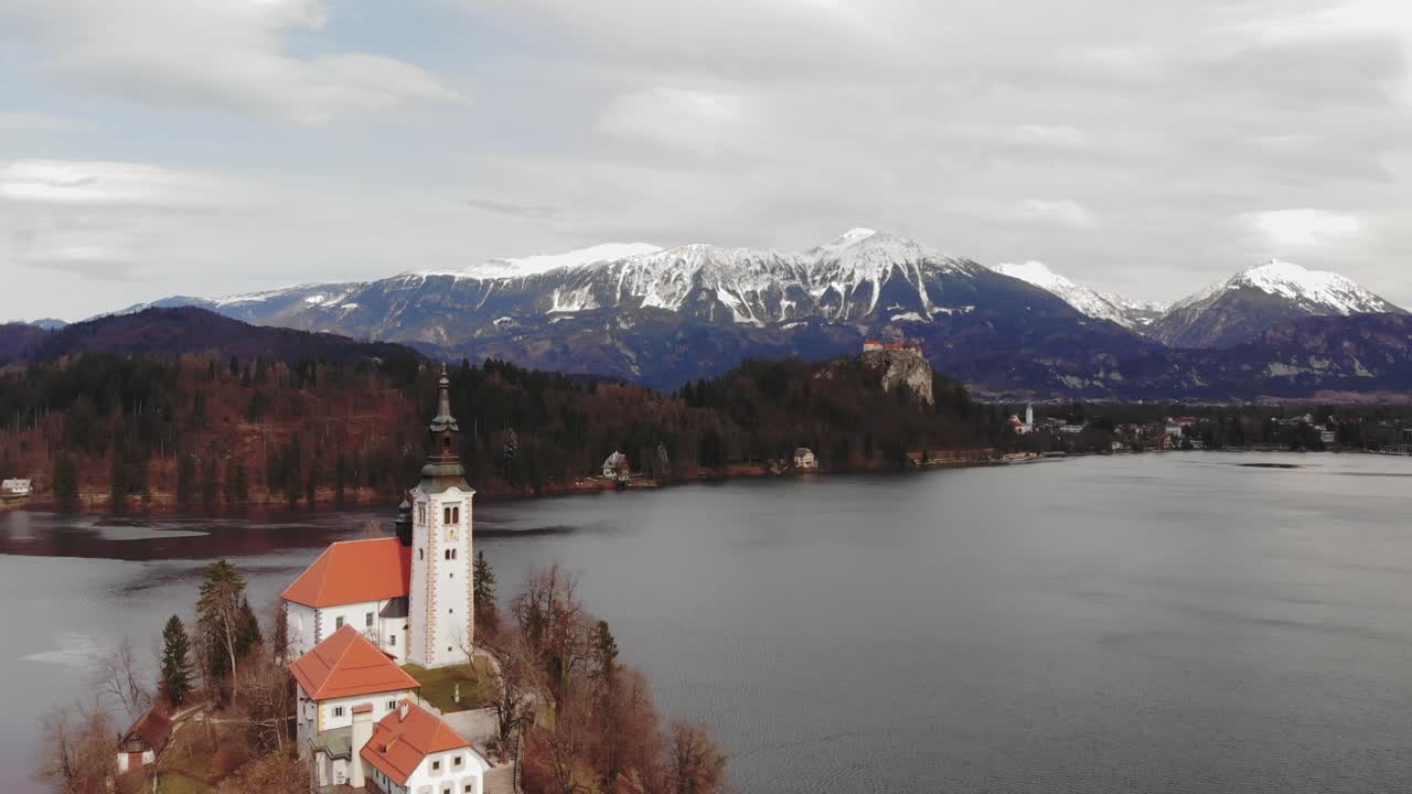 Dronevlucht over oude kerk met berglandschap met indrukwekkende sneeuwtoppen op een bewolkte koude dag, Bledmeer Slovenië