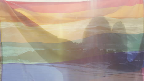 animation of sunset coast over happy diverse gay male couple holding rainbow flag