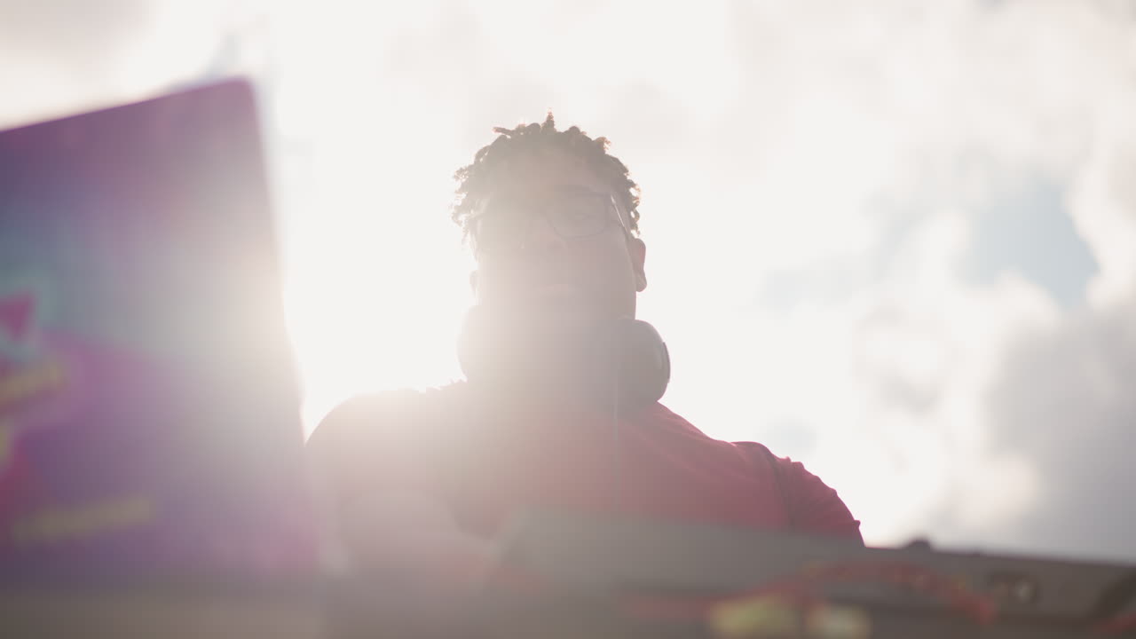 DJ negro mezclando en una mesa al aire libre con laptop y controlador, silueta a contraluz bajo el sol brillante con las manos en los platos, pegatina colorida en la laptop, ambiente de festival enérgico y ritmo concentrado en un set diurno.