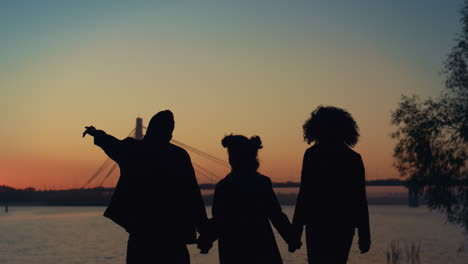 parents silhouette holding hands going to river shore with daughter sunset view.