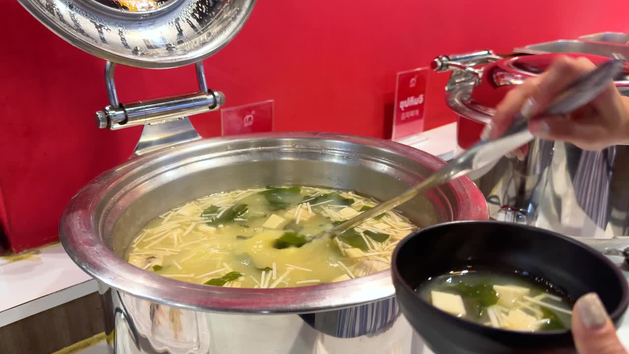 A person serves miso soup from a buffet pot into a bowl at a Korean BBQ restaurant in Bangkok