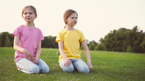 Dos-Lindas-Hermanitas-Hablando-Juntas,-Mirando-Alrededor-Y-Señalando-Algo-Mientras-Se-Arrodillan-Sobre-El-Césped-En-El-Parque-1