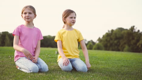 dos lindas hermanitas hablando juntas, mirando alrededor y señalando algo mientras se arrodillan sobre el césped en el parque 1