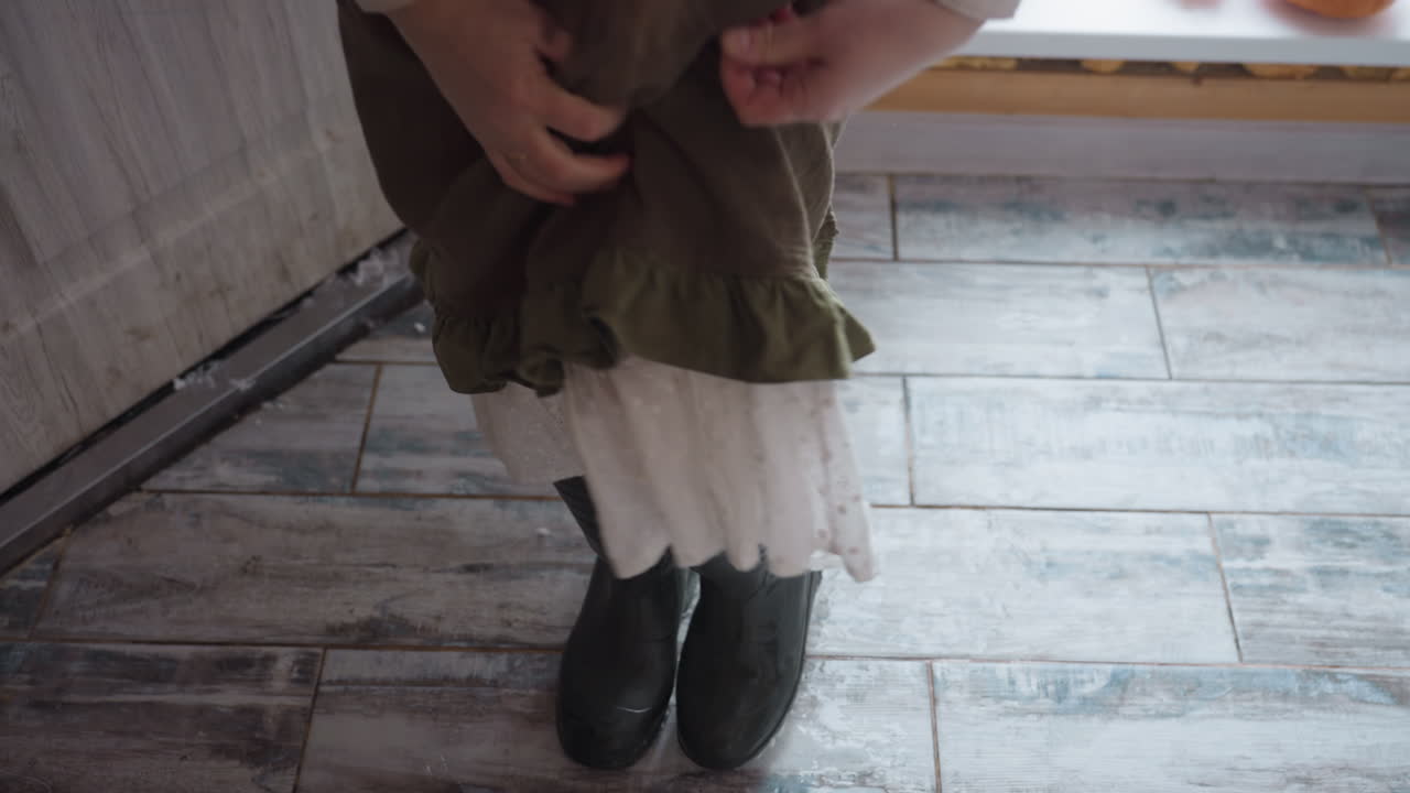 Gardener bending to remove sturdy snow boots while stepping through doorway onto rustic tiled floor, dress ruffles brushing leg, droplets pooling inside threshold, winter light casting soft glow