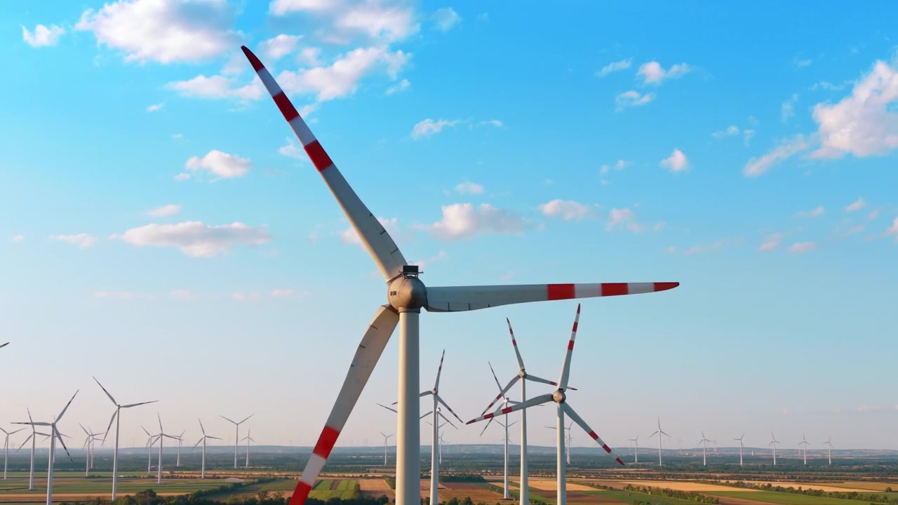 Grande turbina eolica sopra un paesaggio agricolo. Pale a strisce rosse dominano una vista aerea di campi e file di turbine distanti