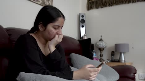 British-Asian-Female-Teenager-Sat-On-Sofa,-Scrolling-Through-Mobile-Phone