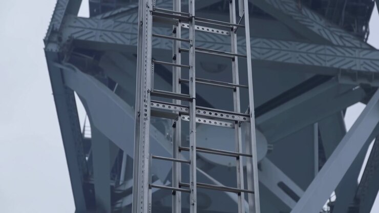Escada vertical de alumínio montada em uma grande superestrutura de aço pintado vista de um ponto de vista ascendente em ângulo baixo, a escada em primeiro plano ocupa a faixa vertical central