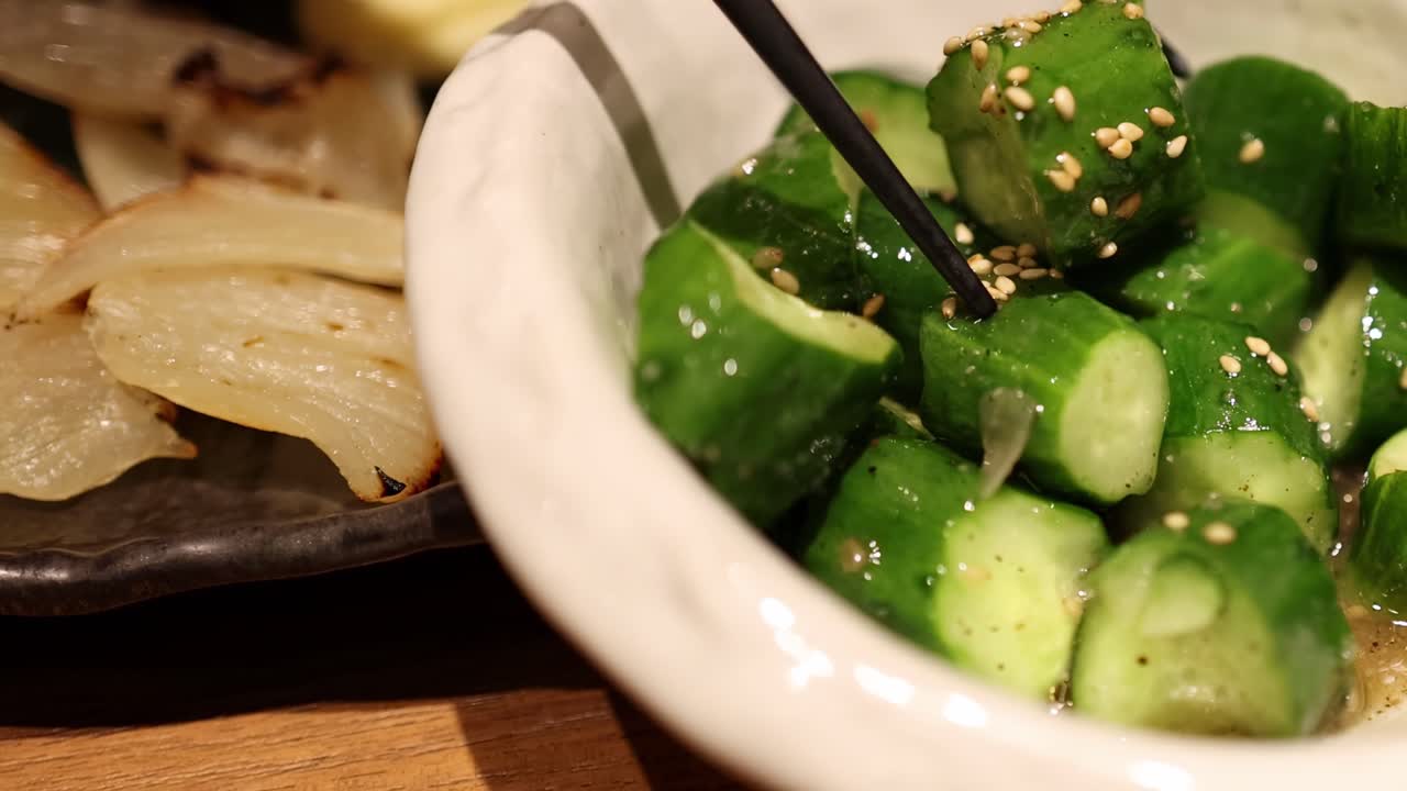 Close-up of cucumber salad garnished with sesame seeds, accompanied by grilled onions on a wooden table.