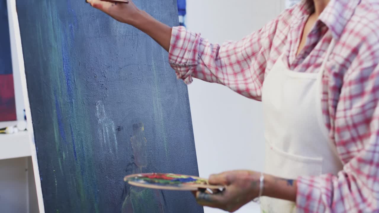 sección media de una artista biracial en delantal pintando en lienzo en el estudio, en cámara lenta