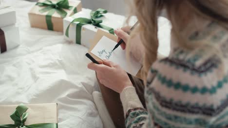 Top-view-of-caucasian-woman-sitting-on-bed-and-writing-Christmas-card-for-wrapped-book