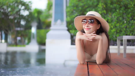 Mientras-Está-Tumbada-Boca-Abajo-En-Una-Terraza-De-Madera-Que-Rodea-Una-Gran-Piscina-De-Un-Complejo,-Una-Mujer-Joven-Con-Un-Sombrero-Para-El-Sol-Y-Gafas-De-Sol-Apoyada-En-Sus-Manos-Sonríe-Mientras-Mira-A-Través-De-La-Propiedad-Del-Hotel