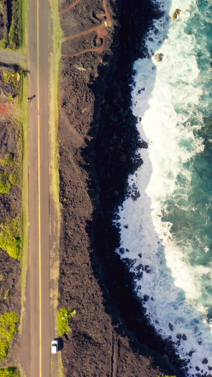 Auto percorrono un'autostrada costiera fiancheggiata da una vegetazione tropicale lussureggiante e scura roccia vulcanica mentre onde spumeggianti si infrangono sotto, mostrando lo straordinario contrasto delle Hawaii tra il design umano e la natura selvaggia dell'isola