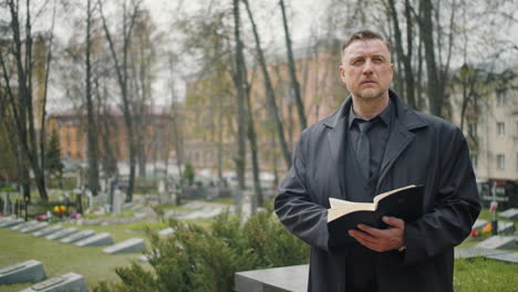 man in black raincoat looking at the horizon in a graveyard and reading a bible 1