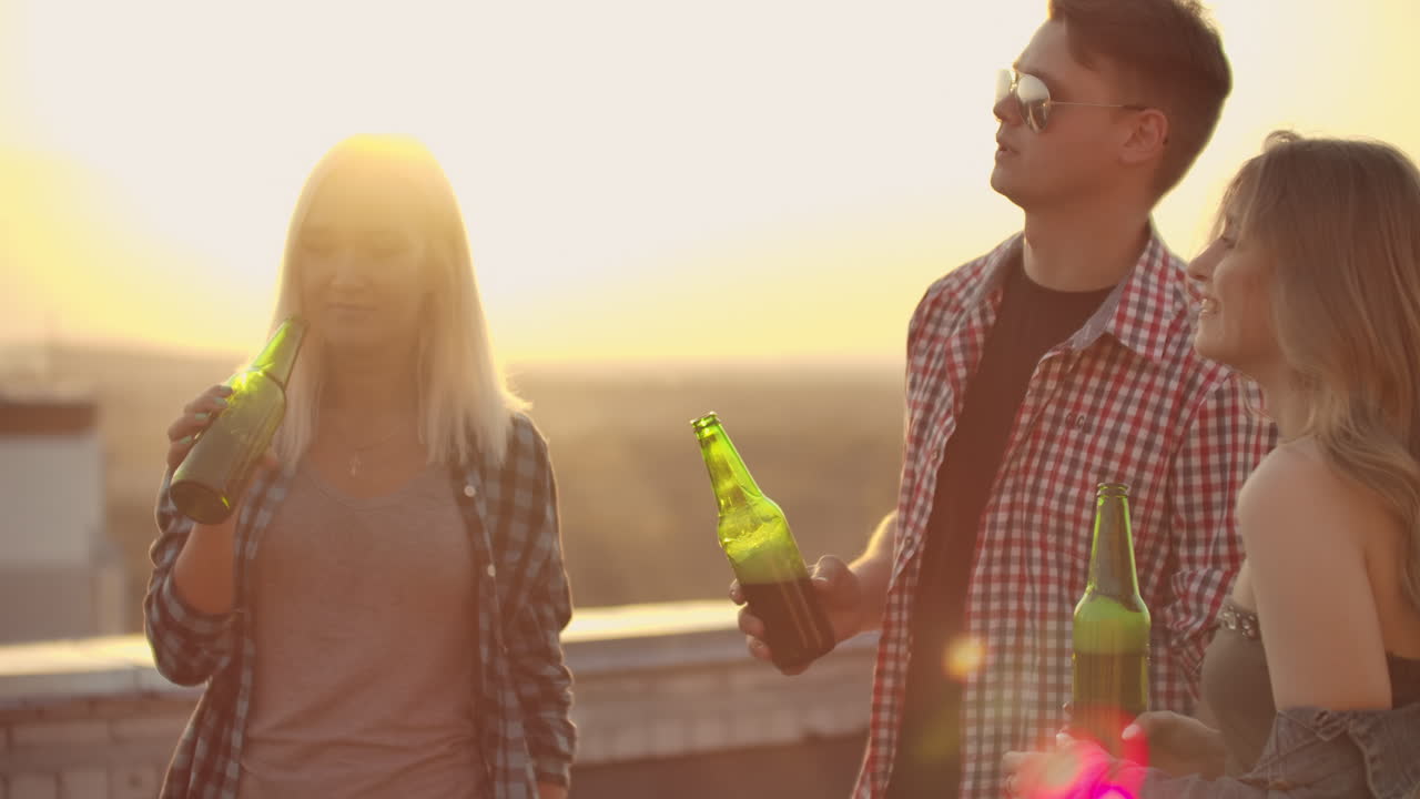 tres chicas y tres chicos disfrutando del tiempo en el techo. sonríen y se comunican entre sí. beben cerveza de botellas verdes y se divierten en las camisas a cuadros.