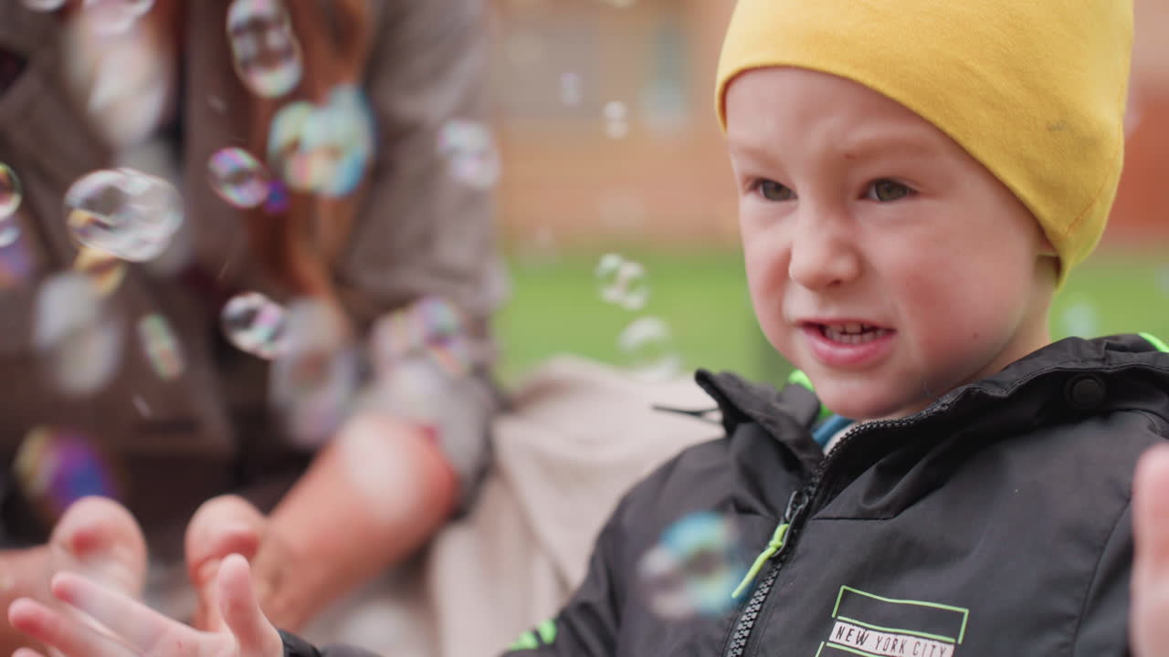 Close up of boy in yellow beanie watching floating soap bubbles, playful focus on face while adult nearby encourages gentle exploration, joyful outdoor moment during courtyard play