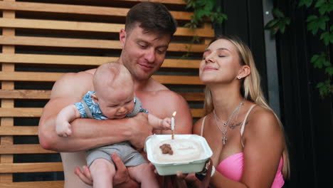 family of three celebrating kid's month anniversary with a cake. happy parents are blowing a candle and kid is starting to cry.