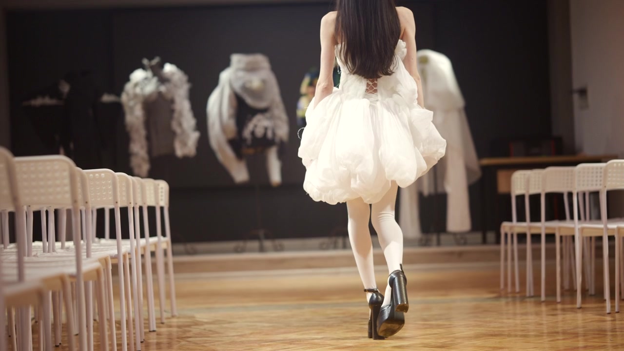 Primo piano di una modella che cammina su una passerella di legno, indossando un voluminoso abito bianco con tessuto a strati e tacchi alti neri.