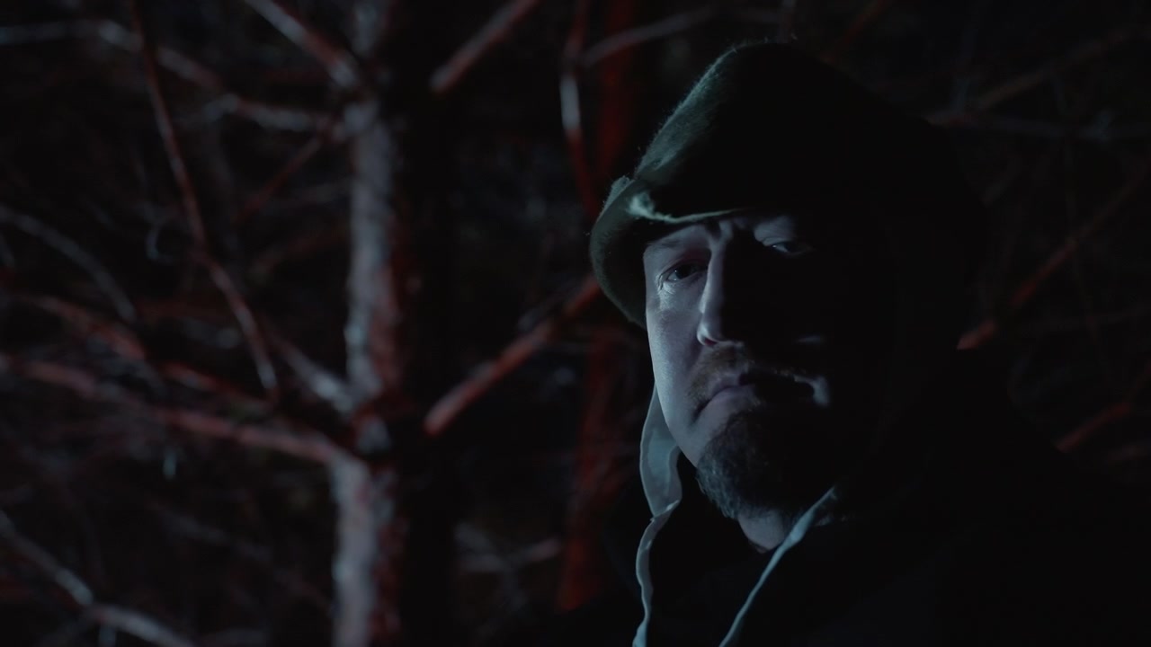 Ritratto notturno ravvicinato di un uomo incappucciato che indossa un berretto con visiera e un cappotto pesante, in piedi accanto a rami di alberi spogli, con il viso parzialmente illuminato da una luce chiave blu fredda e una sottile luce rossa di contorno, creando un'atmosfera tesa e solitaria.