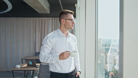 young confident businessman standing in light office. man in white shirt drinking coffee looking into panoramic window.