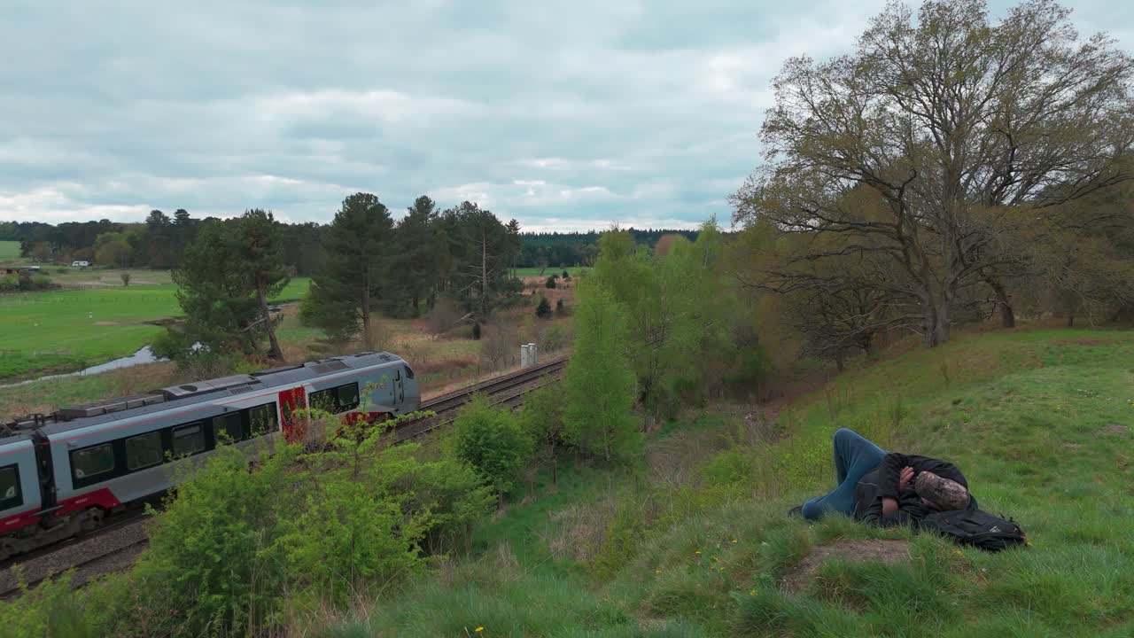 Lone person resting by train tracks with a passing train and lush nature backdrop