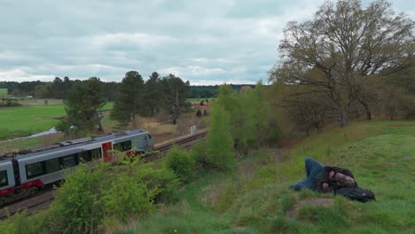 lone person resting by train tracks with a passing train and lush nature backdrop