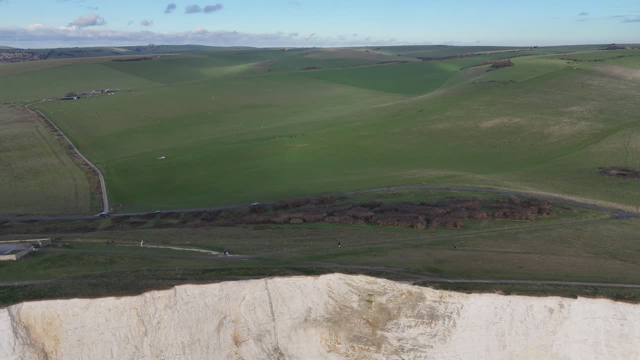 White chalk cliffs rise above rocky coast near winding road, red lantern Beachy Head Lighthouse stands at water’s edge under open sky, dramatic aerial view of East Sussex coastline