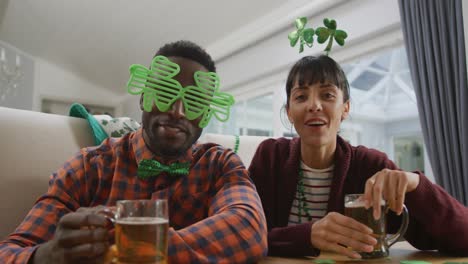 portrait of happy diverse couple with beer wearing clover shape items and having video call