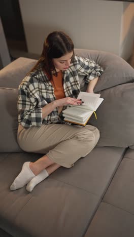 vertical video from above of a confident and calm brunette girl in a checkered shirt and an orange t-shirt sitting on a gray sofa and reading a book at home in a modern apartment