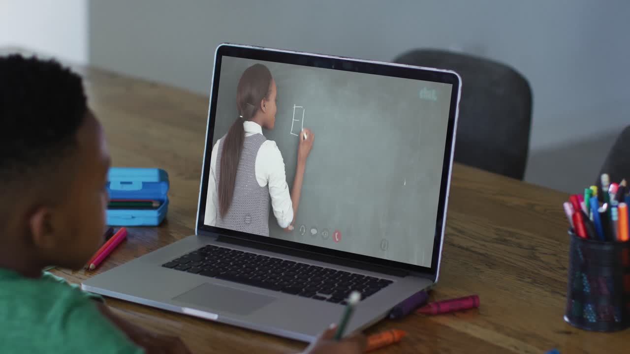 niño afroamericano haciendo la tarea mientras tiene una videollamada con una maestra en una computadora portátil en casa