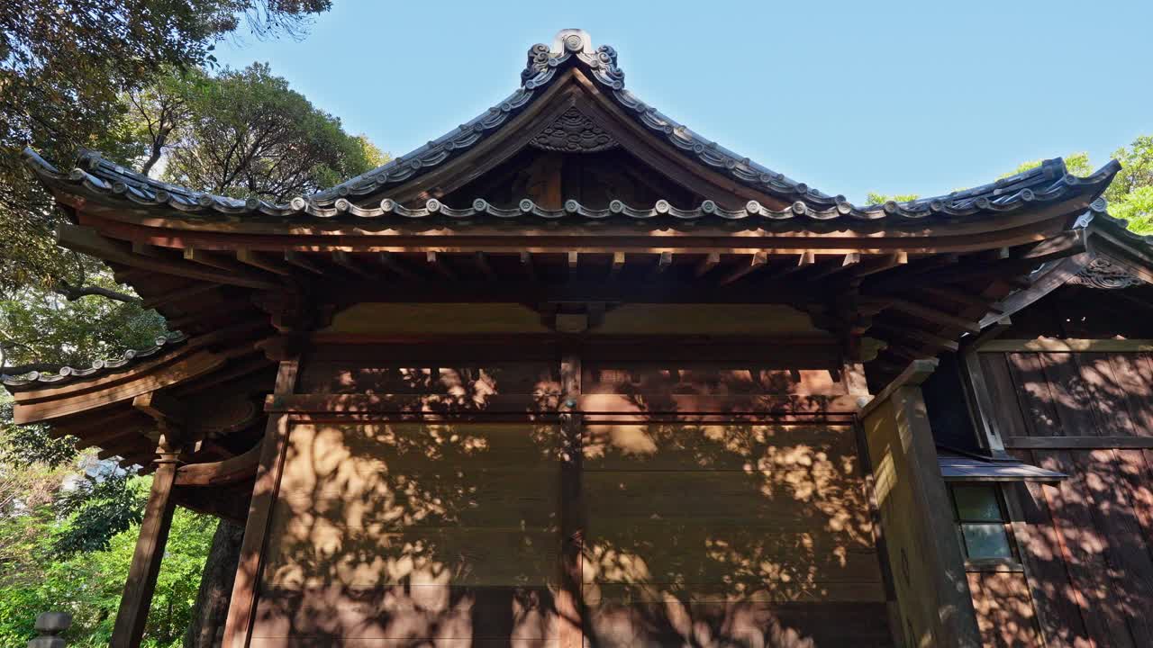 The ornate wooden roof of a traditional Japanese shrine is highlighted by sunlight and dappled with shadows from lush trees.