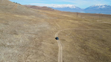 Truck-driving-along-a-dirt-road-in-a-dry-pasture-revealing-a-lake-and-snow-capped-mountains-in-the-distance
